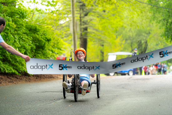 A smiling athlete using a handcycle crosses the finish line at the adaptX 5K, wearing an orange helmet and glasses, surrounded by a green tree-lined road.