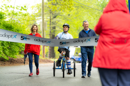 A smiling participant using a handcycle crosses the finish line at the adaptX 5K, flanked by two supporters, as someone in a red coat holds the ribbon.