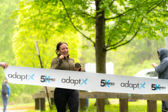 Alt Text: A smiling runner wearing headphones crosses the adaptX 5K finish line on a rainy day, as volunteers hold up the tape beneath green trees.