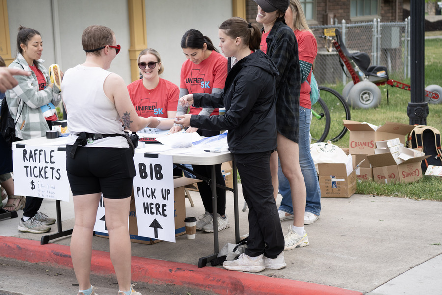 Volunteers in red "Unified 5K" shirts assist participants at a bib pick-up and raffle ticket table during an outdoor event, with boxes and a specialized wheelchair in the background.