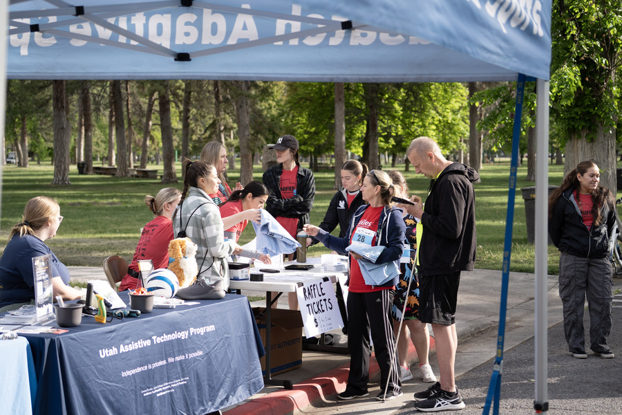 Volunteers and participants gather under a canopy at an outdoor event, visiting the Utah Assistive Technology Program booth and a raffle ticket table.