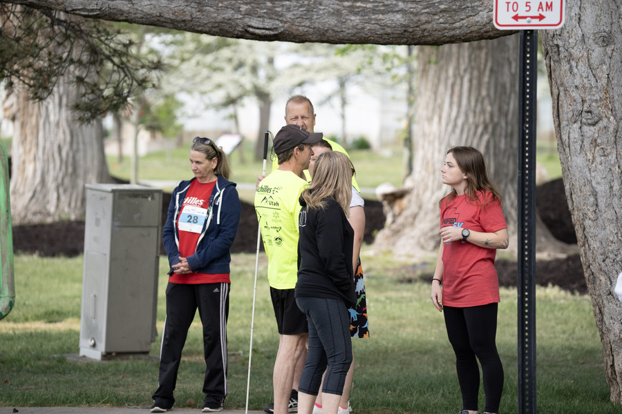 A small group of people talk under a large tree at an outdoor event, including volunteers in red Unified 5K shirts and a man in a bright yellow Achilles International shirt holding a white cane.